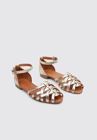 Sandalias planas metálicas doradas con correas en el tobillo, que presentan un diseño trenzado en la parte delantera. Plantilla marrón suave y tacón bajo. Hebilla ajustable.