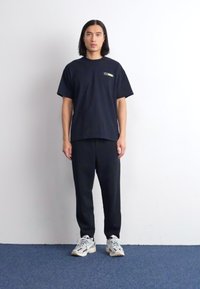 Schwarzes Kurzarm-T-Shirt mit kleinem Logo, kombiniert mit schwarzen Hosen und grauen Sneakers. Glattes Material, lockere Passform und einfaches Design.