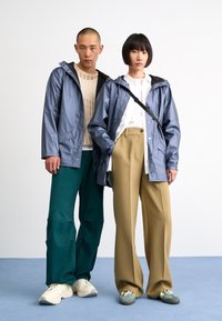 Deux mannequins portent des vestes d'extérieur : l'une en bleu brillant, l'autre en chemise blanc cassé. Les pantalons sont verts et beiges, avec des chaussures contrastantes.