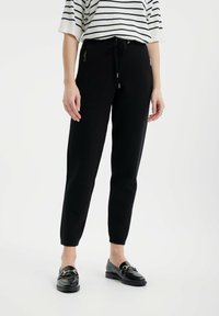 Schwarze, schmal zulaufende Jogginghosen mit einem Kordelzugbund, ausgestattet mit Reißverschlusstaschen an den Seiten und einer glatten, weichen Textur. Kombiniert mit schwarzen Slippern.