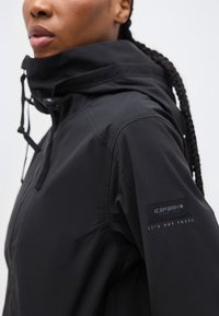 Άτομο που φοράει μαύρο μπουφάν με κουκούλα, με ένα αυτοκόλλητο που γράφει "ICEPEAK IT'S OUT THERE" στο επάνω μέρος του μανικιού, κοιτάζει προς τα αριστερά με πλεξούδες στα μαλλιά.