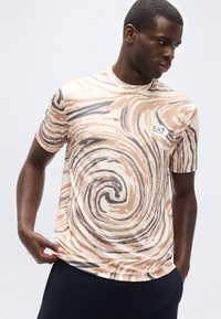 Άνδρας που κοιτάζει προς τα πλάγια, φοράει κοντομάνικο πουκάμισο με μπεζ και καφέ σχέδιο στροβίλου και λογότυπο EA7, κρατώντας το κάτω μέρος του πουκαμίσου, με απλό ανοιχτό φόντο.