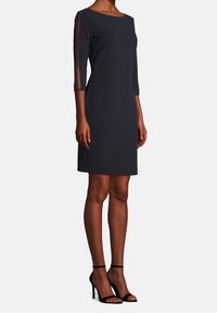 Abito blu navy scuro con maniche trasparenti a tre quarti, design aderente e taglio al ginocchio. Abbinato a sandali neri con tacco alto e cinturini.