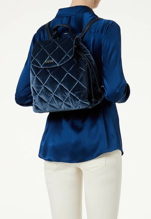 Laivastonsininen samettireppu tikatuskuviolla ja kullanvärisillä logokoristeilla, jossa on yläkahva ja säädettävät olkahihnat.