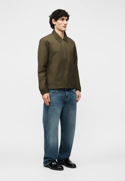 Homme debout portant une veste zippée vert olive, un jean bleu ample et des chaussures noires, devant un fond clair uni.