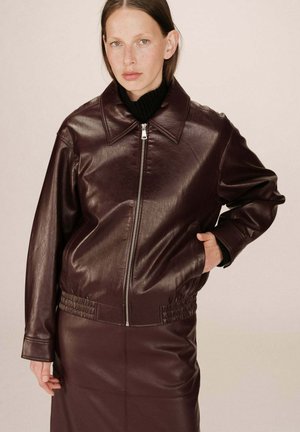 Veste en cuir bordeaux avec fermeture éclair à l'avant, col pointu et ourlet côtelé. Le modèle porte un col roulé noir. Texture lisse, design minimal.