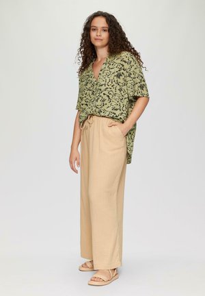 Kurzärmlige grüne Bluse mit schwarzem Blumenmuster, kombiniert mit lockeren beigen Hosen und hellbraunen Sandalen. Lässiger Schnitt, lässiger Stil.