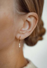 Goldene Creolen mit zwei kleinen weißen Perlen und einem goldenen Akzent, die daran hängen, werden an einem Nahaufnahme eines Ohrs präsentiert.