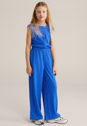 Combinaison bleue texturée avec un corsage boutonné et une taille élastique. Comprend un pantalon à jambes larges et des manches courtes. Associée à des baskets argentées.