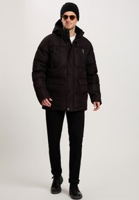 Czarna kurtka puchowa z kapturem, z zamykanymi kieszeniami na piersi i guzikami, noszona z czarnymi spodniami i butami, tworząc warstwowy wygląd.