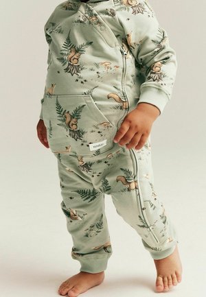 Taapero, joka pukeutunut vaaleanvihreisiin, orava- ja metsäkuvioituihin housuihin ja takkiin, seisoo paljain jaloin yksiväristä taustaa vasten.