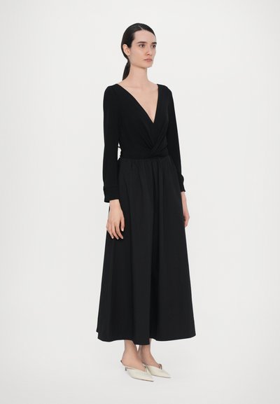 Ilga juoda suknelė ilgomis rankovėmis, su giliais V formos iškirpte, susuktos bodžio detalės, surinkta juosmens sritis ir plačiai teka sijonas. Derinama su šviesios spalvos slip-on bateliais.