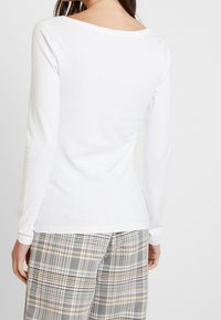 Bílá košile s dlouhým rukávem a širokým výstřihem, hladká textura a přiléhavý střih, doplněná kostkovanými kalhotami v neutrálních tónech.