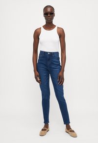 Blå høy midje jeans laget av denim, med en tettsittende silhuett, klassisk fem-lomme design, og knappestolpe. Brukt med en hvit tank topp.