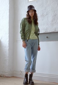 Casaco bomber verde por cima de uma blusa verde clara, jeans azuis claros enrolados e botas de tornozelo pretas. O modelo usa um chapéu preto estilo bucket.