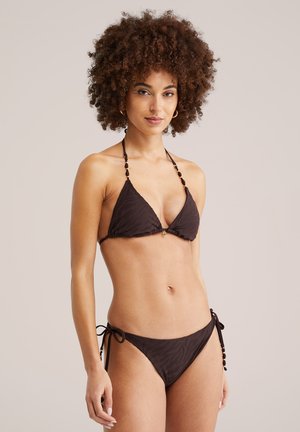 Femeie cu păr creț, purtând un bikini maro închis cu model, stând în picioare pe un fundal simplu bej deschis.