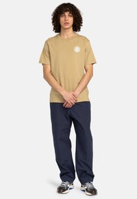 Beige kortærmet t-shirt med rund hals og et hvidt logo på venstre bryst, parret med marineblå løse bukser og mønstrede sneakers.