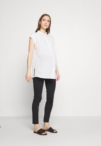 Blusa branca de manga curta, com colarinho e corte reto, com fendas laterais, combinada com jeans pretos de corte slim e sandálias pretas tipo slide.
