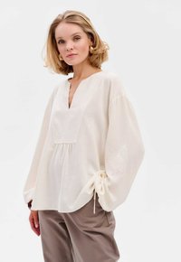 Cremfarbene Leinenbluse mit tiefem V-Ausschnitt, weiten Ärmeln und elastischen Bündchen. Verziert mit zarter Stickerei an den Ärmeln. Kombiniert mit taupefarbenen Hosen.