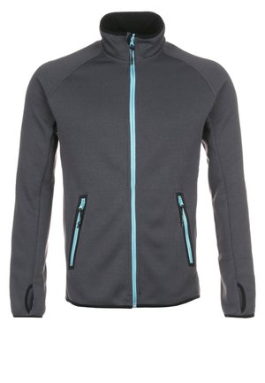 Homme portant une veste zippée gris foncé sur un t-shirt bleu, un pantalon noir et des chaussures de randonnée noires, debout devant un fond uni.