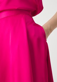 Fuksia kjole med en glatt satinvare, en tettsittende midje med belte og flytende stoff som fremhever mykhet og livlig farge.