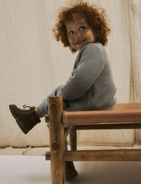 Bambino seduto su una panchina di legno, indossando un maglione grigio a texture e pantaloni grigi, abbinati a stivaletti marroni con lacci. Lo sfondo è neutro.