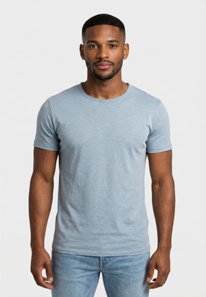 Uomo con capelli corti e barba che indossa una T-shirt a maniche corte azzurro chiaro e jeans blu, in piedi contro uno sfondo chiaro uniforme.