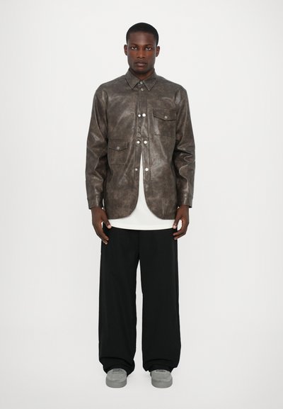 Veste chemise en cuir synthétique brun avec un col boutonné, deux poches poitrine, portée sur un haut blanc, associée à un pantalon noir ample et des chaussures grises.