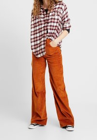 Kariertes Flanellhemd in Weiß, Burgunderrot und Blau; Cord-Hose mit weitem Bein in Orange; schwarze Sneaker mit niedrigem Schaft; sichtbare Vordertaschen.