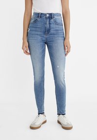 Svetle modré vysoké skinny džíny s mírně opotřebeným vzhledem, v kombinaci s bílými teniskami, na jednobarevném bílém pozadí.