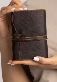 Carnet en cuir marron avec une surface texturée, maintenu par une sangle en cuir plus claire, présentant un design à rabat et des détails de couture visibles.