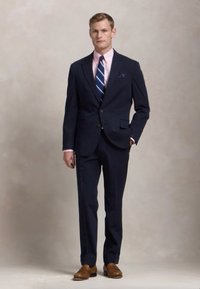 Costum cu dungi navy, cu sacou cu un rând de nasturi, cămașă roz deschis, cravată cu dungi albastre și pantofi din piele maro. Inclus un batiste pentru buzunar.