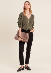 Olivgrüner, gestrickter Reißverschluss-Pullover, schwarze, eng sitzende Jeans, schwarze Slipper mit goldenen Akzenten und eine braune Handtasche mit geflochtenem Griff.