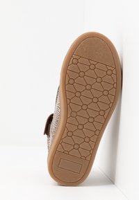 Hnědá podrážka s vzorem, obsahující texturovaný grip, s hexagonálním vzorem a hnědým okrajem. Na boku se nachází uzavření na suchý zip z textilie.