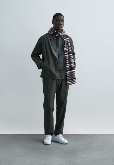 Olivovo zelená bunda a nohavice, modrá košeľa, pletený šál v hnedej, krémovej a fialovej pruhovanej farbe a biele tenisky. Jednoduchý, textúrovaný módny vzhľad.