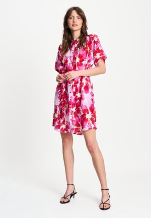 Vrouw staand, gekleed in een kort mouw, knielange roze en rode bloemenjurk met zwarte bandjeshakken tegen een effen witte achtergrond.
