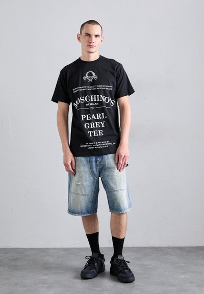 Juodas medvilninis marškinėliai su balta tekstu iš Moschino, derinami su nublukusiomis mėlynomis džinsinėmis šortais. Modelis stovi ant pilko grindinio, avėdamas juodus kojines ir sportinius batelius.
