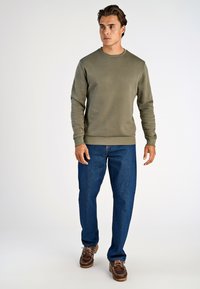 Oliwkowa bluza z okrągłym dekoltem i prążkowanymi mankietami, zestawiona z prostymi niebieskimi dżinsami i brązowymi butami typu boat shoes.