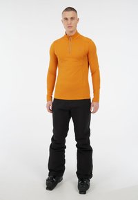 Vyras stovi juodais slidinėjimo batais, juodomis slidinėjimo kelnėmis ir oranžine ilgomis rankovėmis viršutine su ketvirčio užtrauktuku baltame fone.