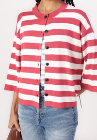 Cardigan a righe rosso e bianco con scollatura rotonda, maniche corte e bottoni neri. Indossato sopra una camicia a righe blu chiaro e abbinato a jeans.