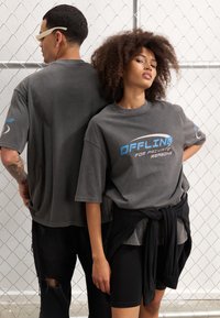 Grå oversized t-shirts med teksten "OFFLINE FOR PRIVATE REASONS" trykt i blå og hvid. Tætsiddende sorte shorts og en bundet sort jakke. Slidte denim bagpå.