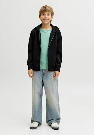 Czarna bluza na suwak na jasnooliwkowy t-shirt, zestawiona z szerokimi niebieskimi dżinsami. Buty białe z czarnymi podeszwami. Prosty, casualowy strój.