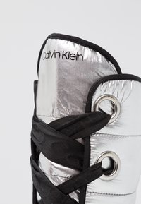 Stříbrná nadýchaná bota s vysokým designem, černými šňůrkami a značkovým logem Calvin Klein. Má lesklý povrch a kovové akcenty.
