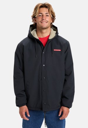 Chaqueta negra con capucha, botones a presión y forro de forro polar beige. Tiene bolsillos laterales y un parche rojo con el logo en el pecho.