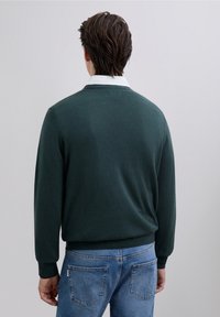 Mörkgrön stickad tröja med rund halsringning och ribbade muddar, bärs över en vit skjorta med krage, kombinerad med ljusblå jeans.