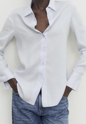 Dark-skinned person wearing an open-collar white button-up shirt and blue jeans with hands tucked into pockets.