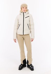 Weißer Puffer-Jacke mit schwarzen Reißverschlüssen und Taschen, kombiniert mit beigen, engen Hosen und schwarzen Skischuhen. Das Modell trägt außerdem eine beige Strickmütze.