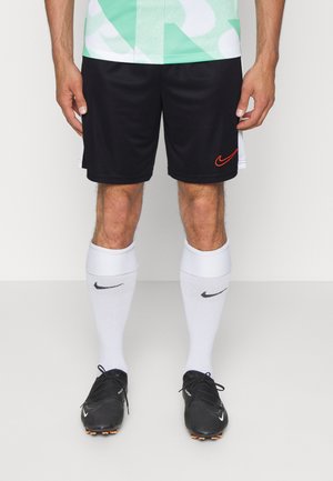 Męskie nogi w czarnych piłkarskich szortach Nike z czerwonym logo, białych, sięgających kolan skarpetach Nike oraz czarnych korkach Nike na jednolitym tle.