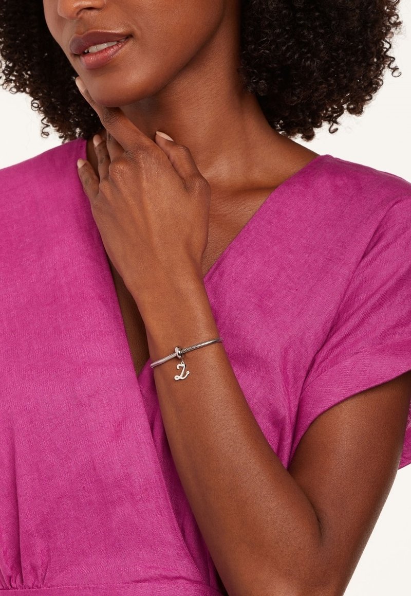 Zilveren armband met een bedel in de vorm van de letter "L," gedragen op een hand tegen een roze gestructureerde stof. Stijlvol ontwerp benadrukt de pols.