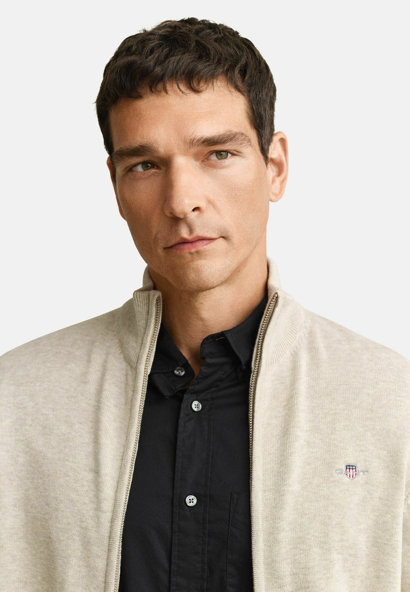 Hombre con cabello corto y oscuro, vistiendo un suéter beige con cremallera sobre una camisa negra abotonada, frente a un fondo blanco liso.